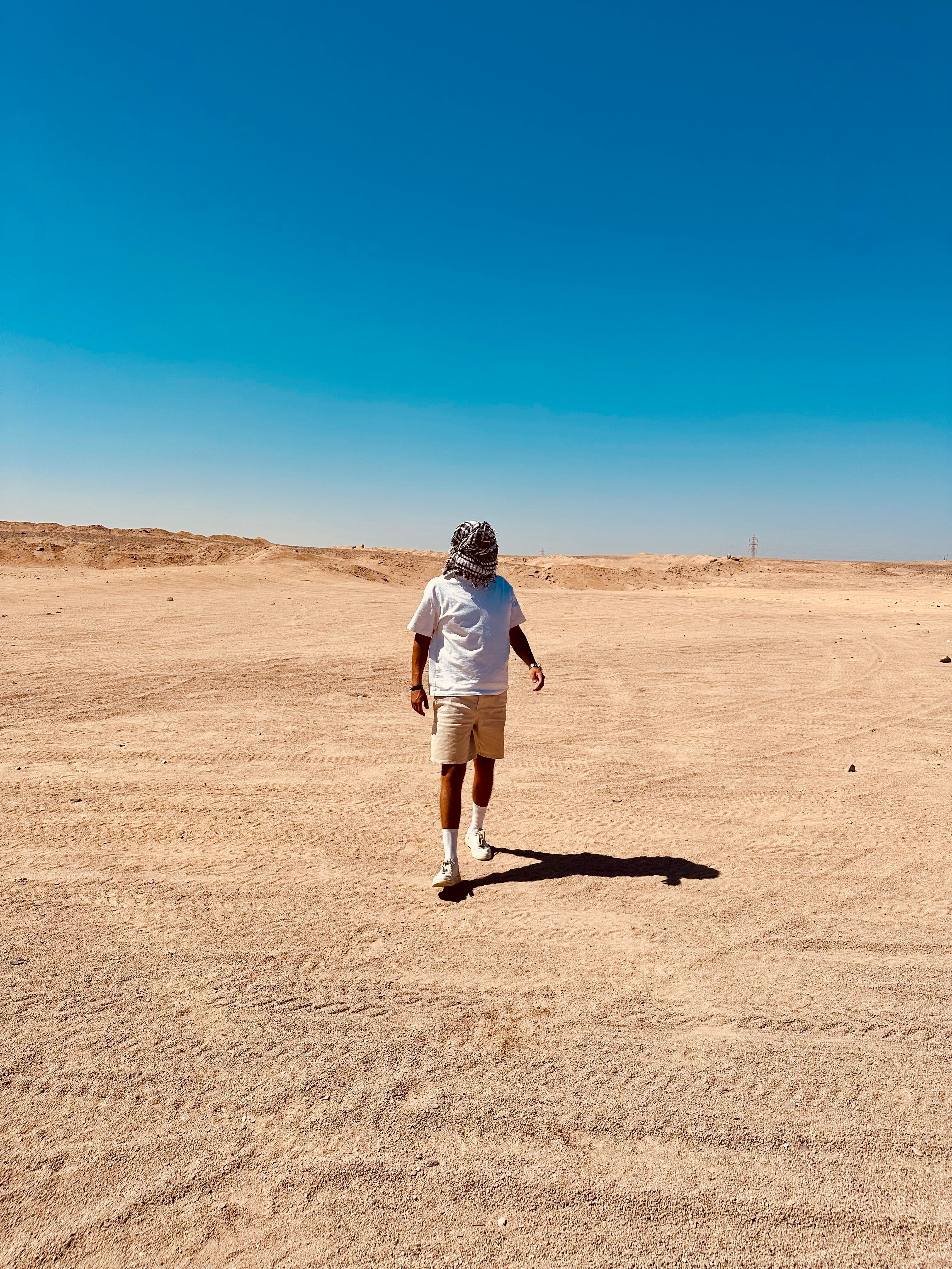 A man walks alone across a vast, sunlit desert landscape under a clear blue sky.