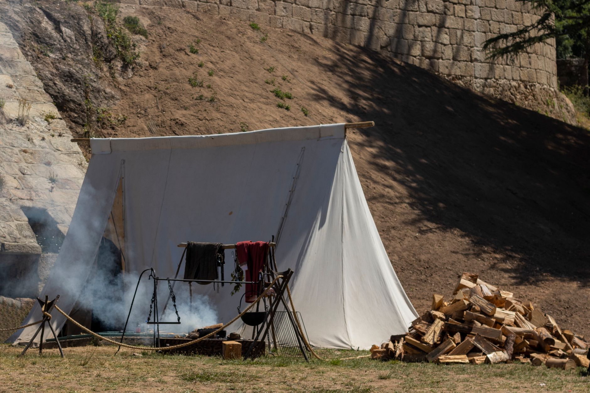 A medieval-style campsite with a white tent, firewood, and smoke outdoors.