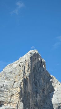 A towering rocky cliff under a clear blue sky with the moon visible at peak.