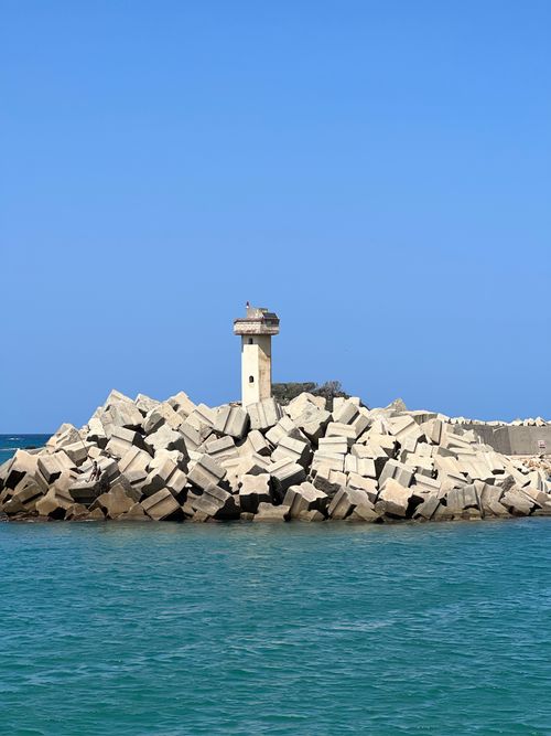 Free Lighthouse stands amidst geometric rocks by the ocean on a sunny day. Stock Photo