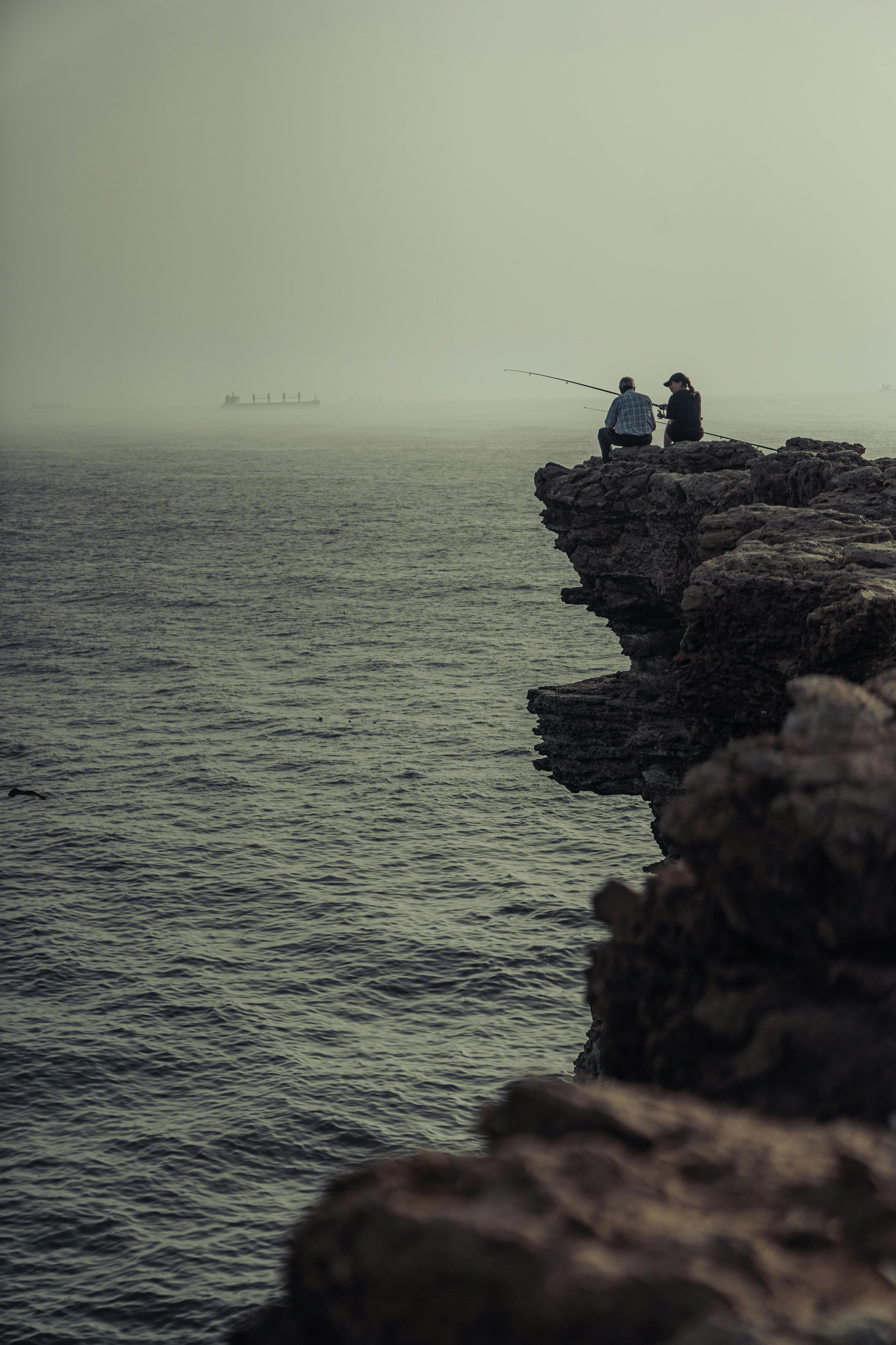 Two fishermen on a rocky cliff in El Jadida, Morocco, casting rods at dusk with foggy seascape surroundings.