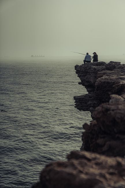 Two fishermen on a rocky cliff in El Jadida, Morocco, casting rods at dusk with foggy seascape surroundings.