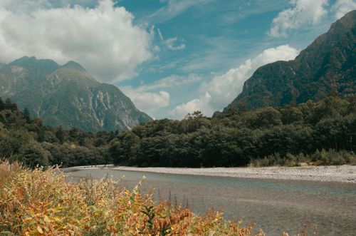 Free Serene river scene with lush green trees and towering mountains under a clear sky. Stock Photo