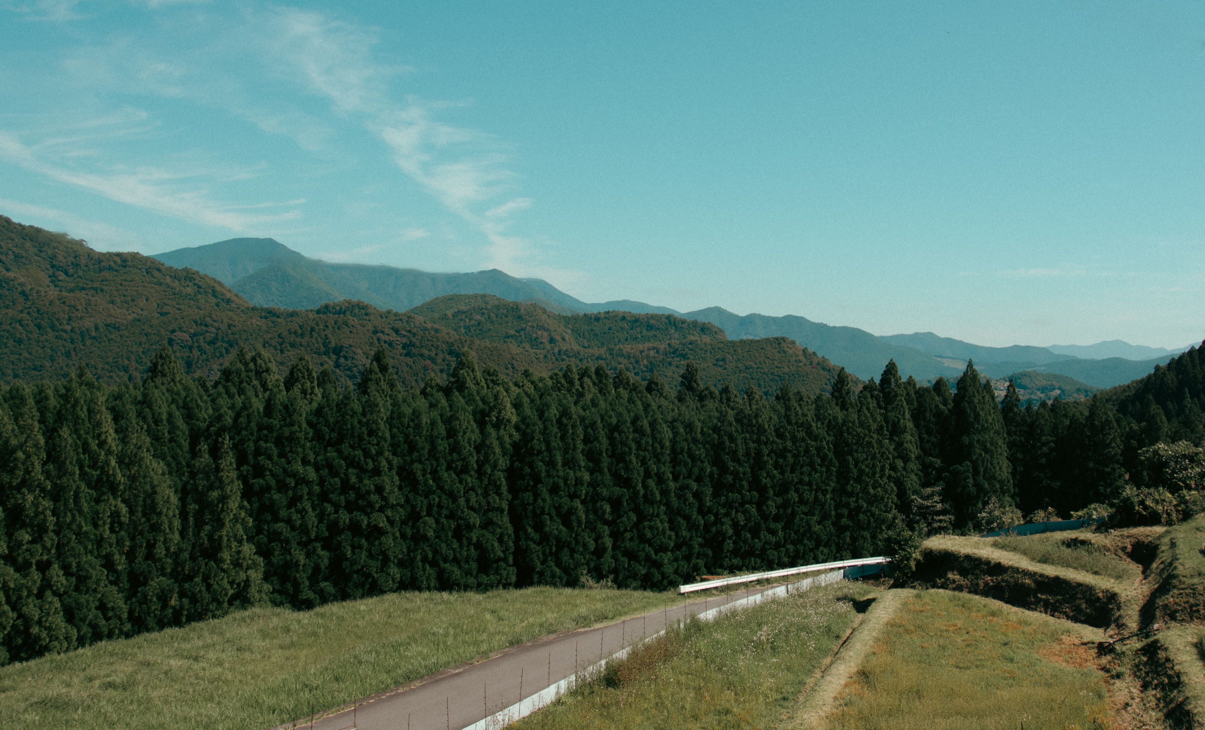 Scenic mountain landscape featuring a serene road flanked by lush forests and rolling hills under a clear blue sky.