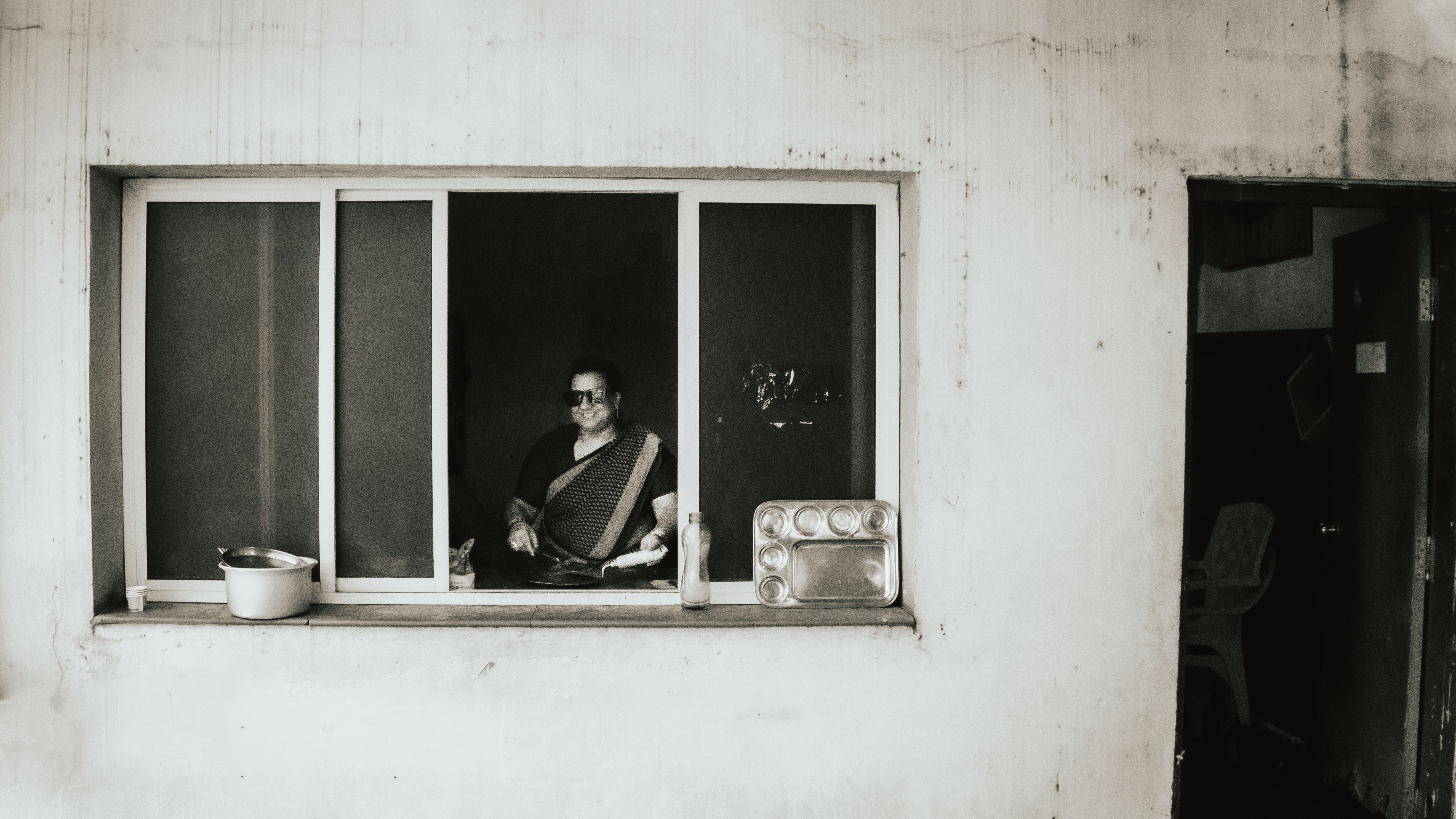 Mulher em saree cozinhando dentro de casa, vista através da janela. Ambiente urbano com utensílios visíveis.