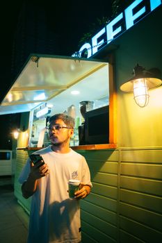 A man holding coffee and phone outside a cafe at night, illuminated by warm lights.