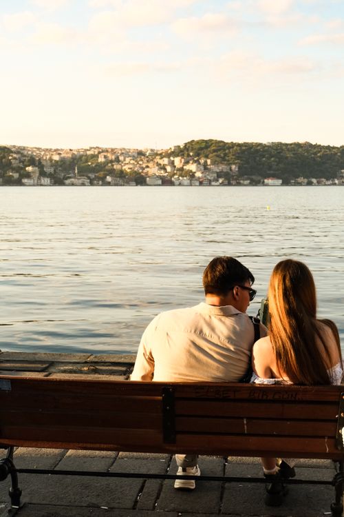 Free A couple sits on a bench enjoying a peaceful sunset by the sea with a cityscape in the distance. Stock Photo