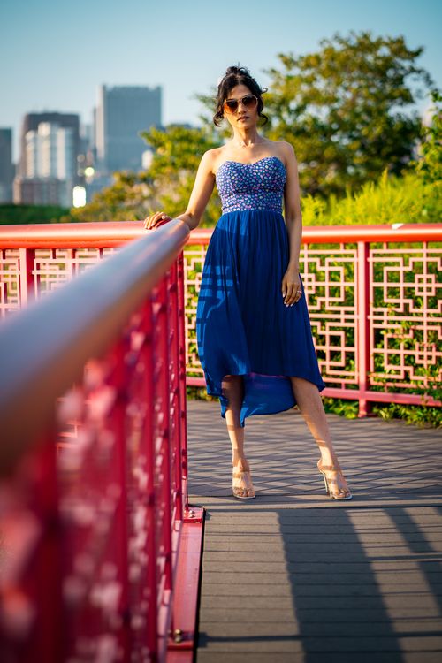 Free Stylish woman in a blue dress poses on a colorful bridge against a city backdrop. Stock Photo