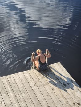 Back view of a woman in swimsuit sitting on a pier by a lake on a sunny day.
