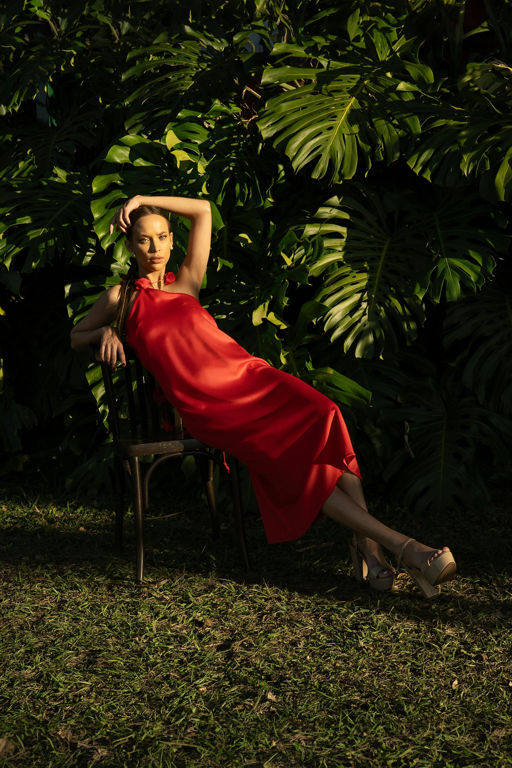 Stylish portrait of a woman in a red dress leaning on a chair in a lush garden.