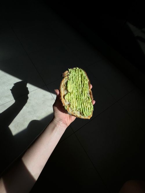 Free A hand holding an avocado toast with sunlight creating shadows on a tiled floor in Amsterdam. Stock Photo
