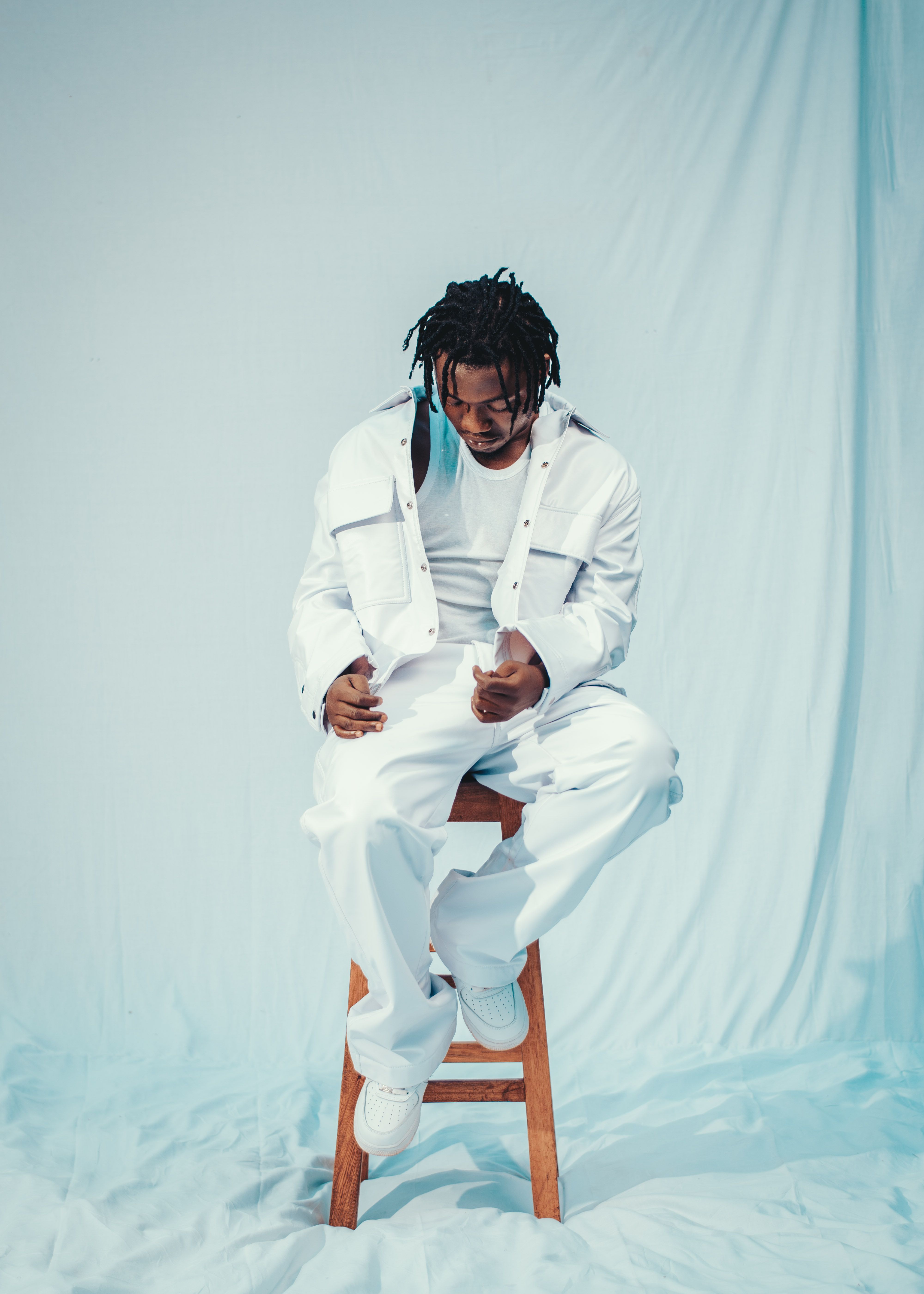 A man in white attire sits on a stool against a light blue backdrop, capturing a serene moment indoors.