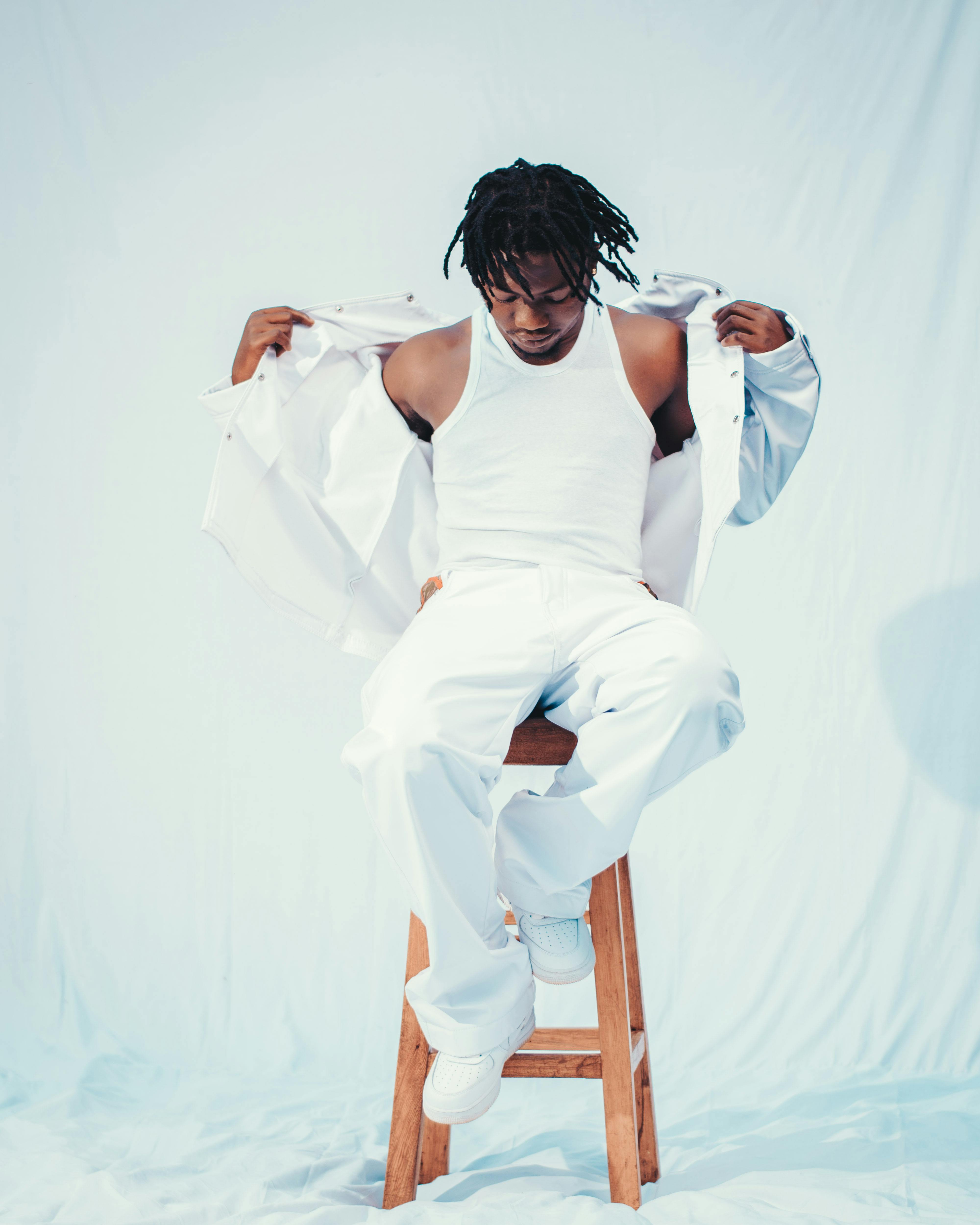 A young man wearing all white clothing sitting confidently on a wooden stool against a light background.