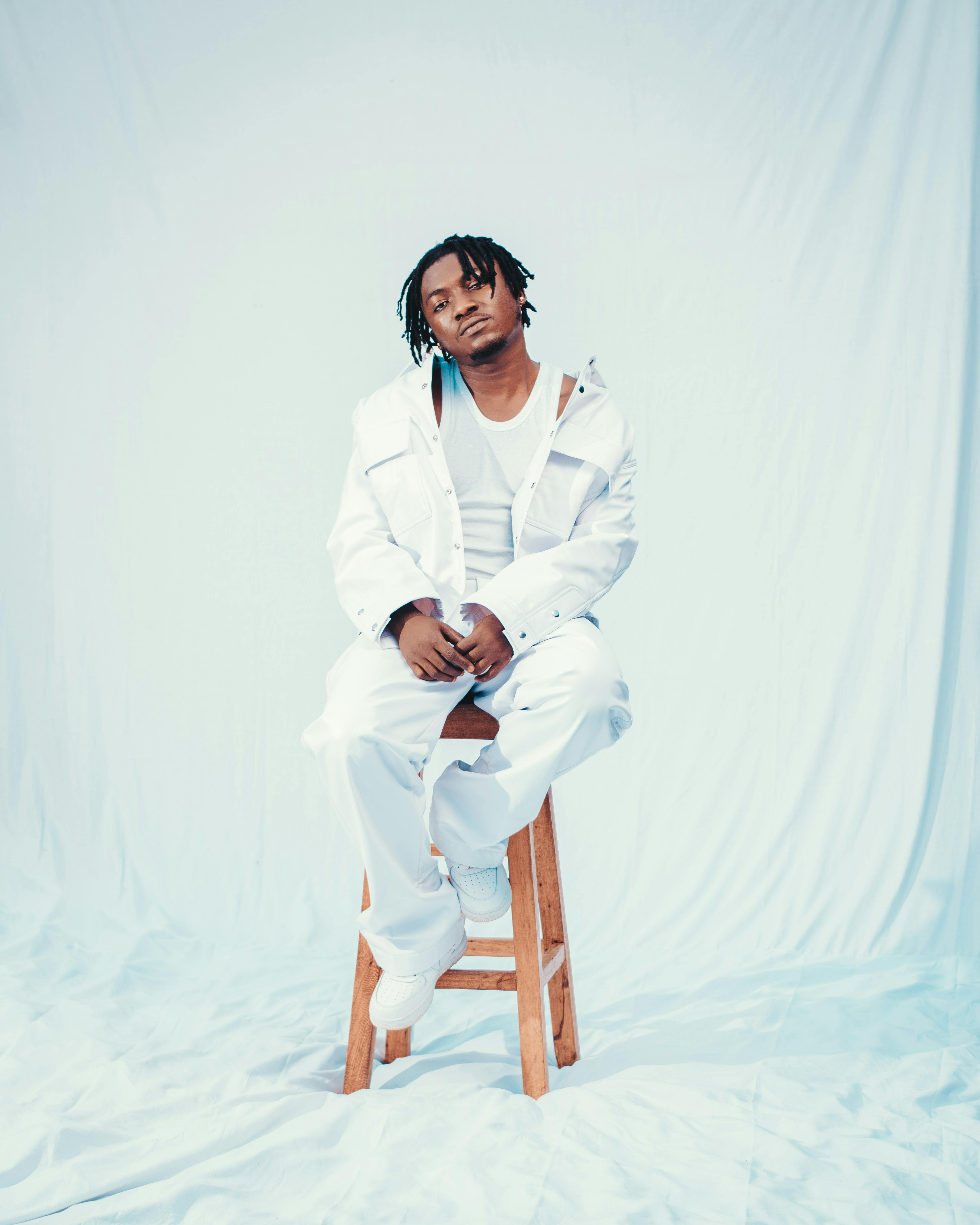 Portrait of a stylish man in white sitting on a wooden stool against a soft backdrop.