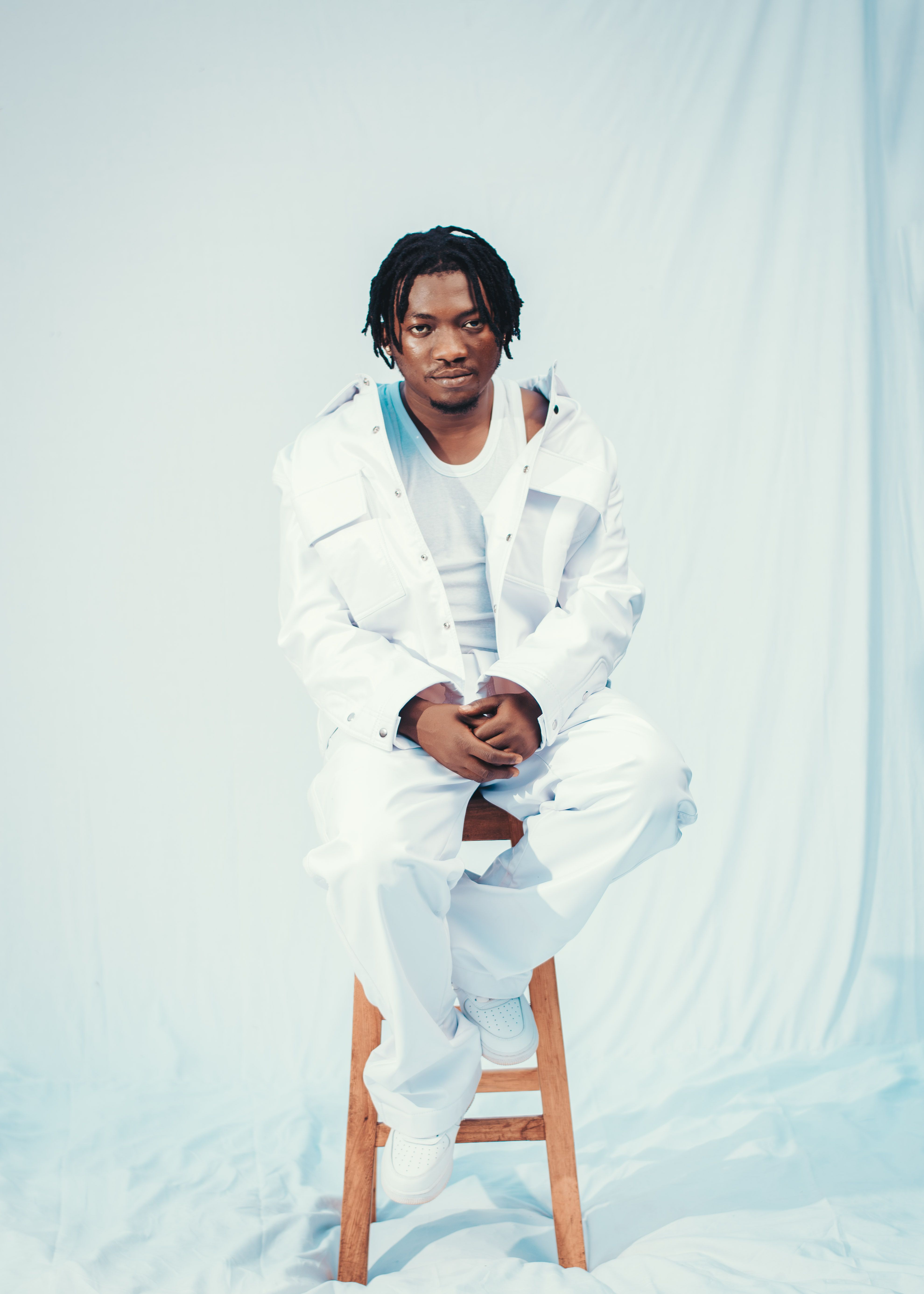 Portrait of a man in white attire sitting on a stool against a white background.