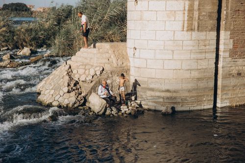 Free Fishermen by the riverbank under a bridge in Al Qanatir, Egypt, enjoying a sunny day. Stock Photo