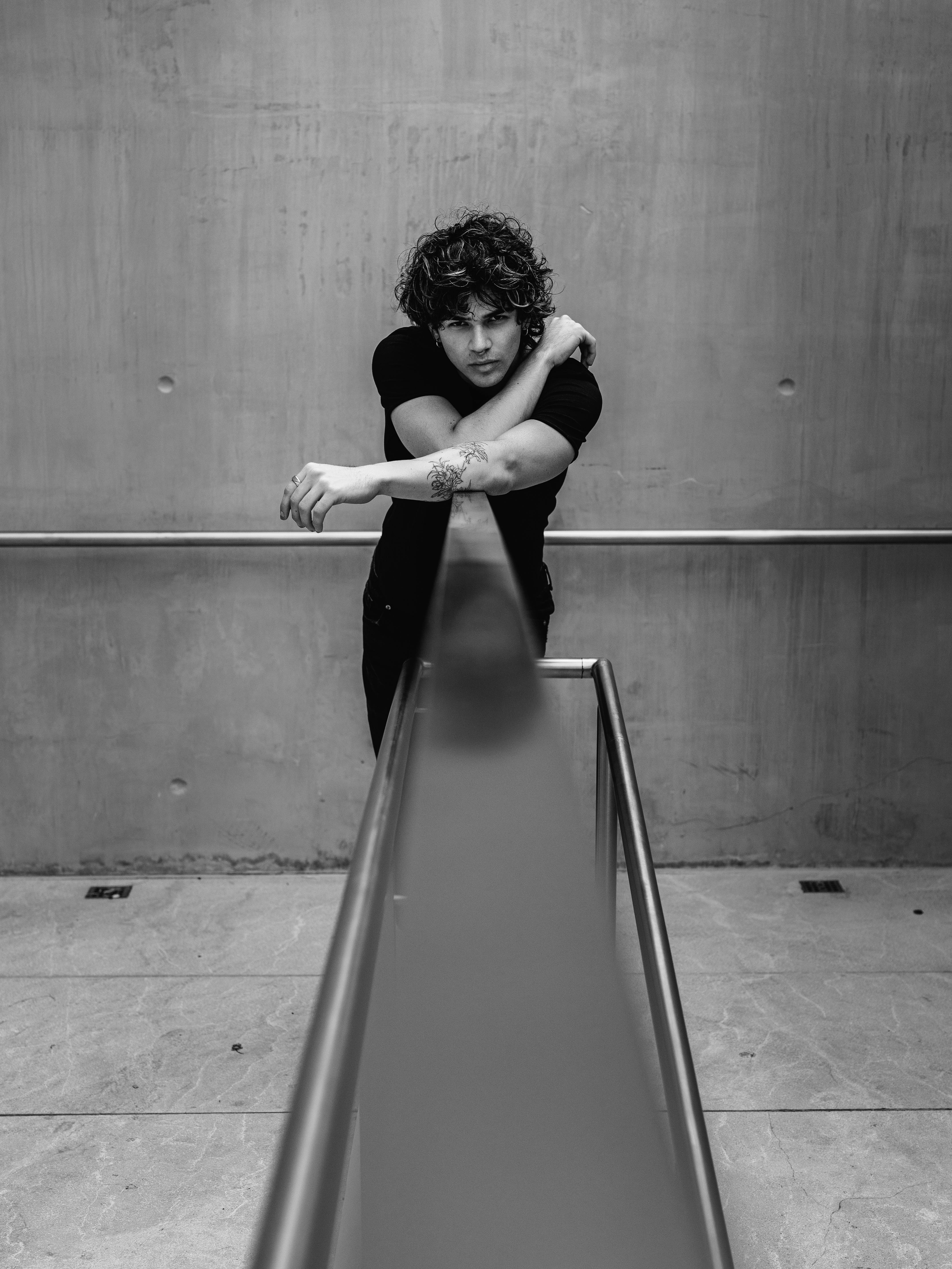 Black and white portrait of a stylish young man leaning over a rail in an urban setting.