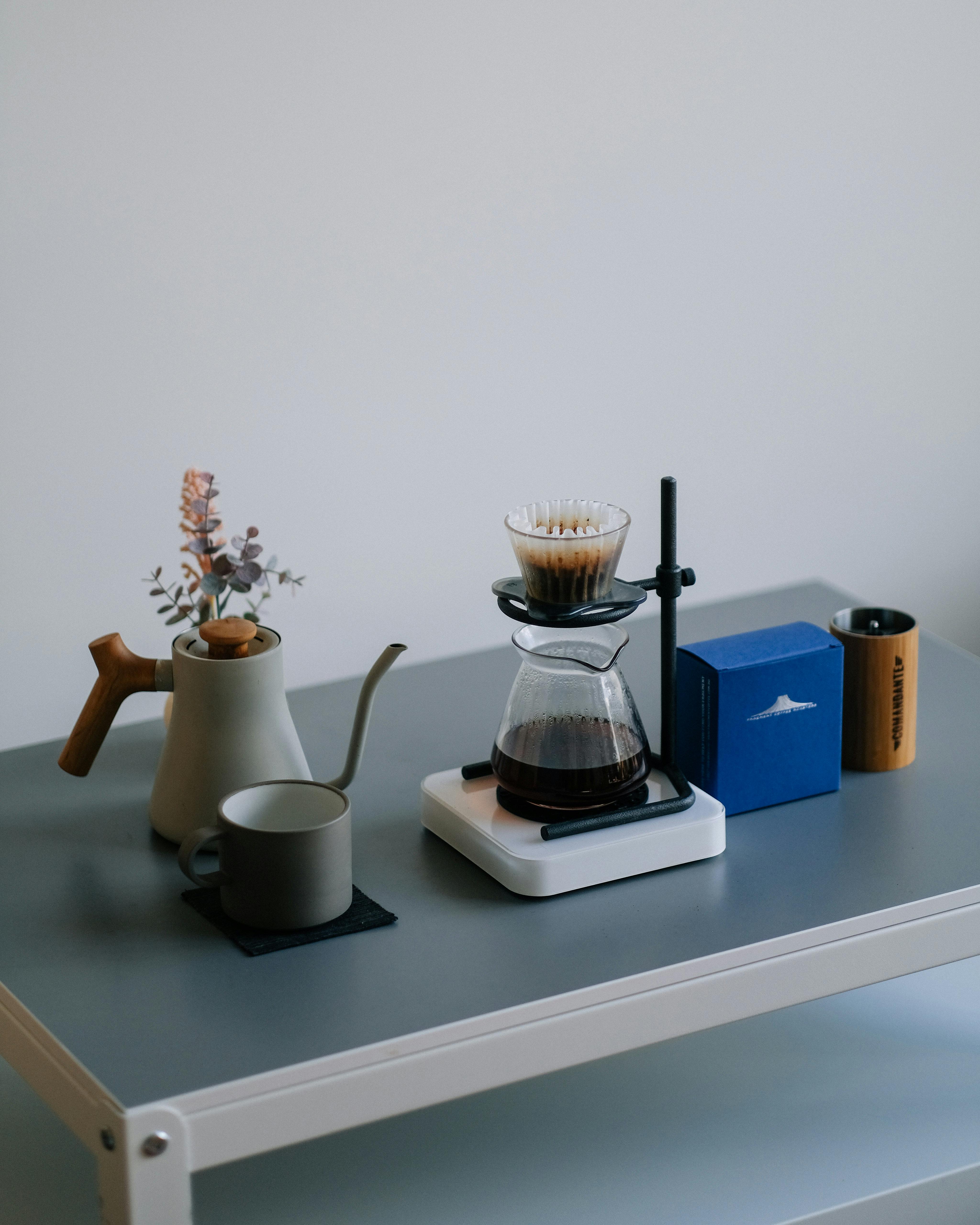A minimalist coffee setup featuring a pour over brewer, ceramic cup, and kettle on a table.