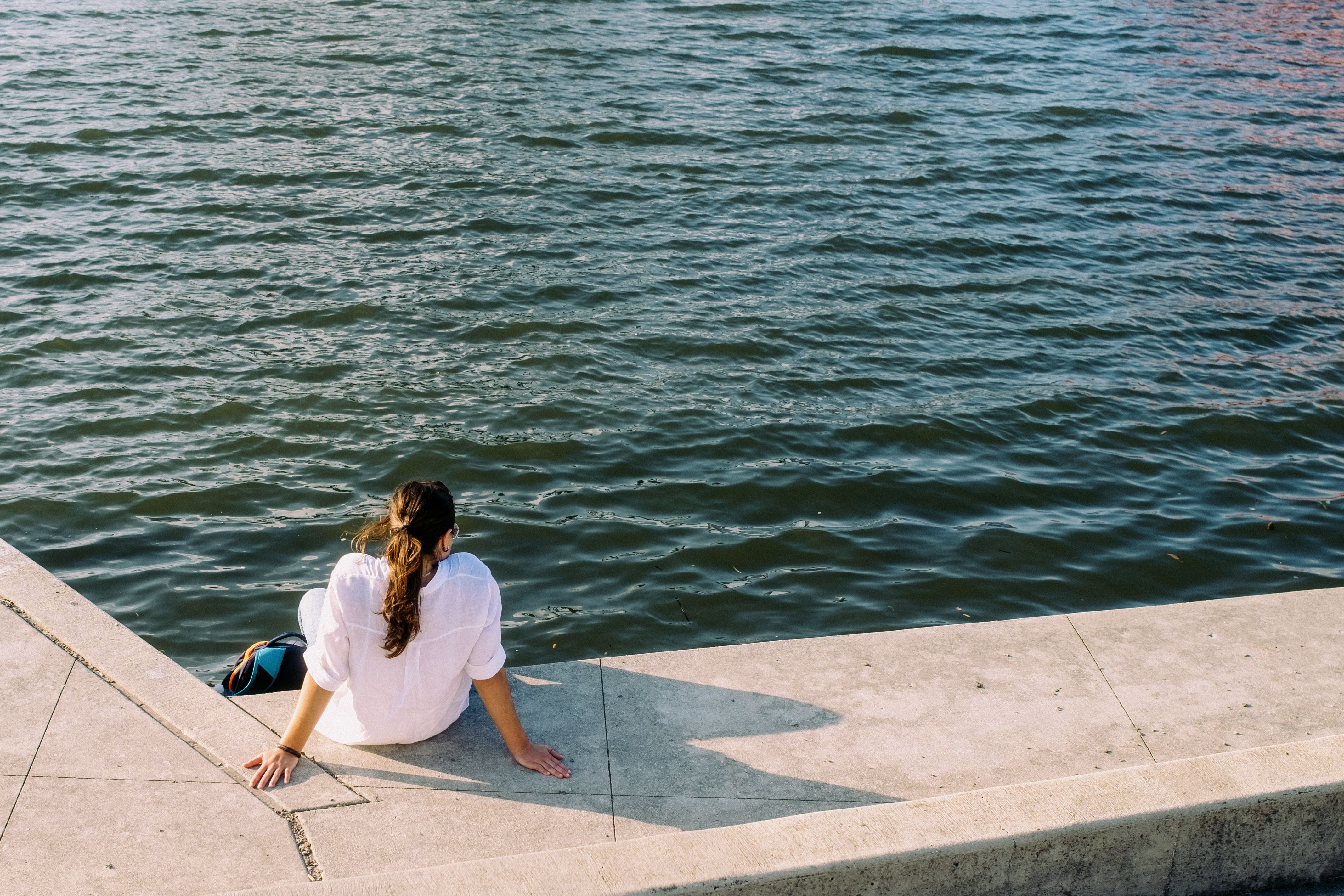 A woman sitting on a concrete ledge looking at the water · Free Stock Photo