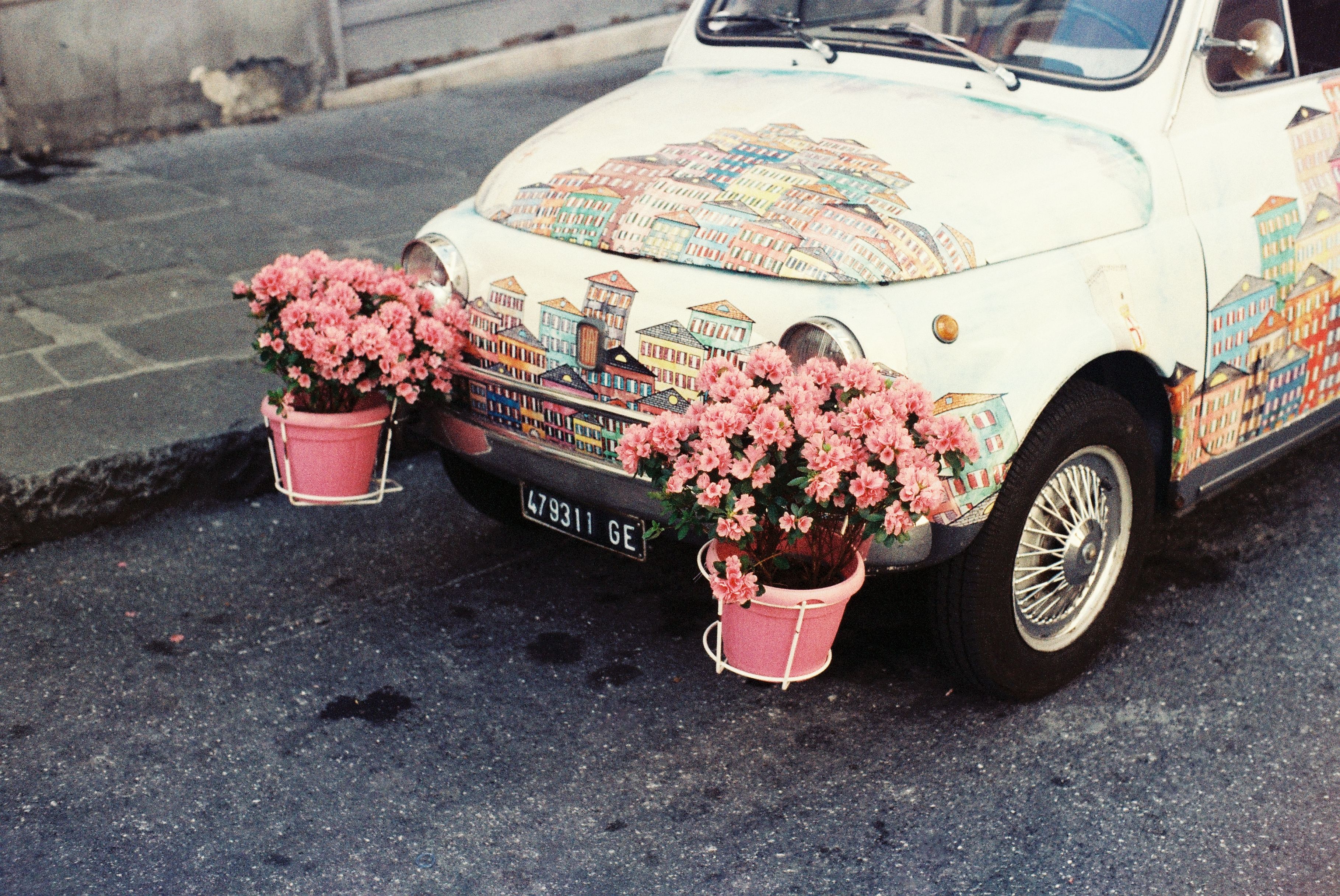 Carro decorado com pintura de cenário urbano e cercado por vasos de flores cor de rosa em uma rua, transmitindo criatividade e charme urbano.