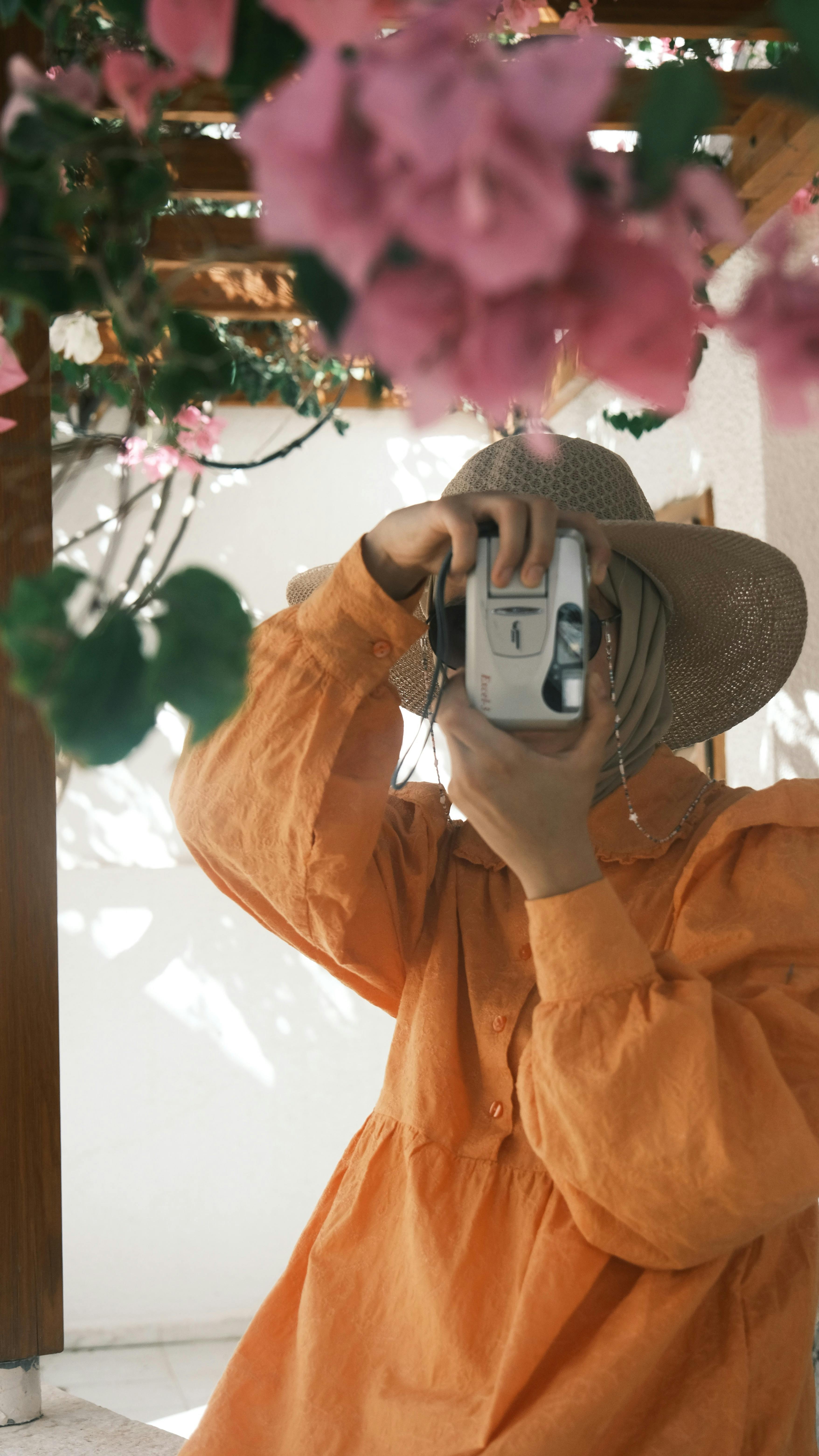 A woman in an orange dress takes a photo surrounded by vibrant flowers outdoors.