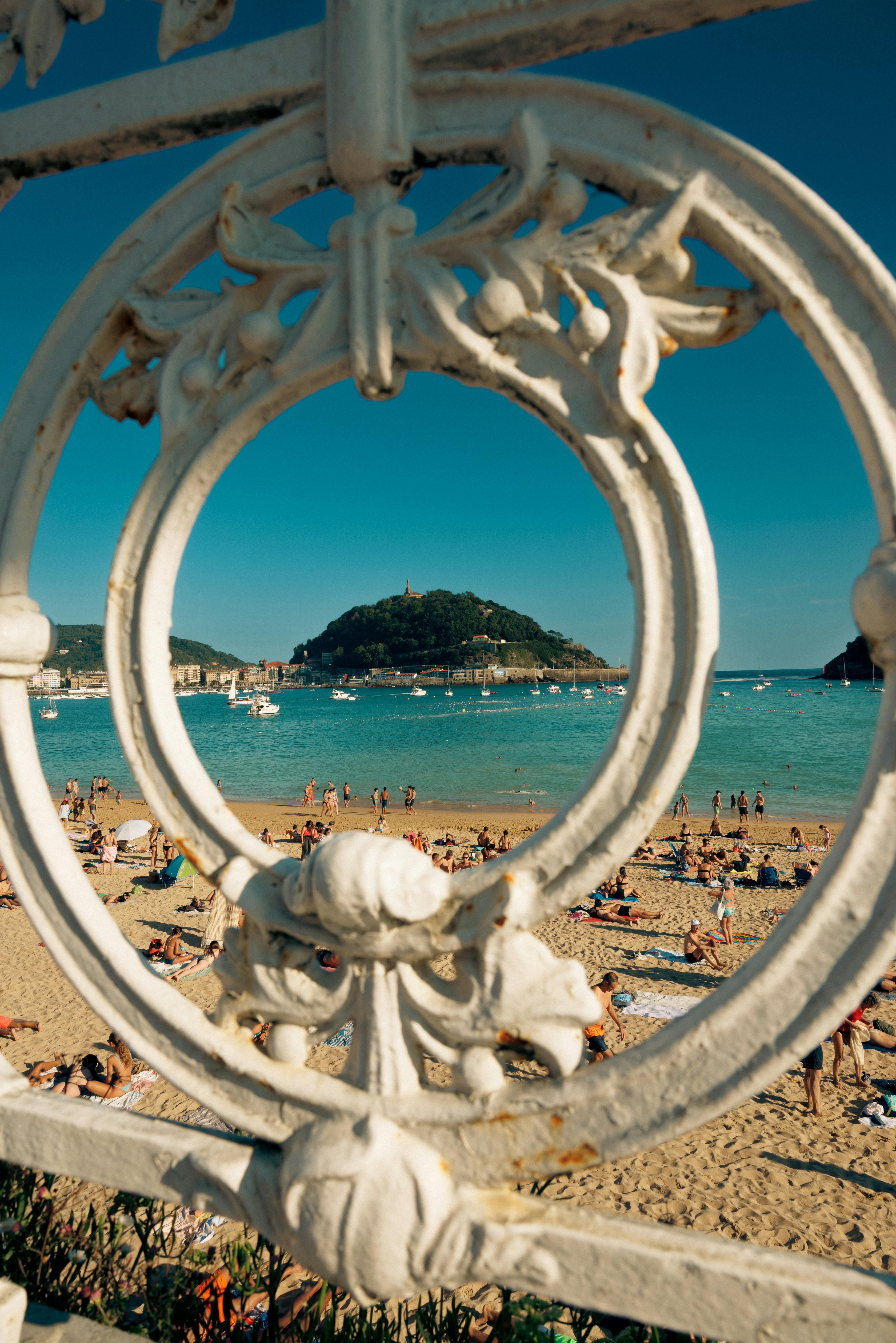 View of crowded beach through ornate ironwork with island in distance.