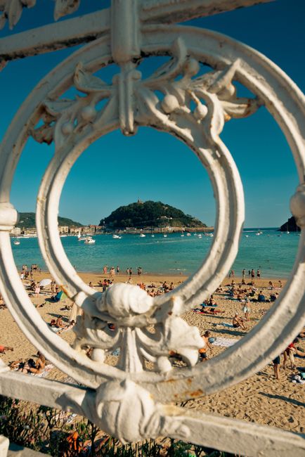 View of crowded beach through ornate ironwork with island in distance.