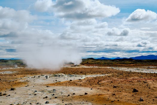 Wide view of a steaming geothermal field with cloudy sky, showcasing natural geothermal activity.
