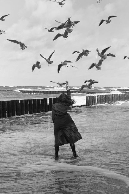 Black and white photo of a woman with seagulls at a windy beach.