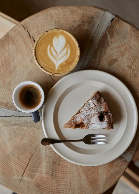 Overhead view of a cappuccino, espresso, and cake on a rustic wooden table.