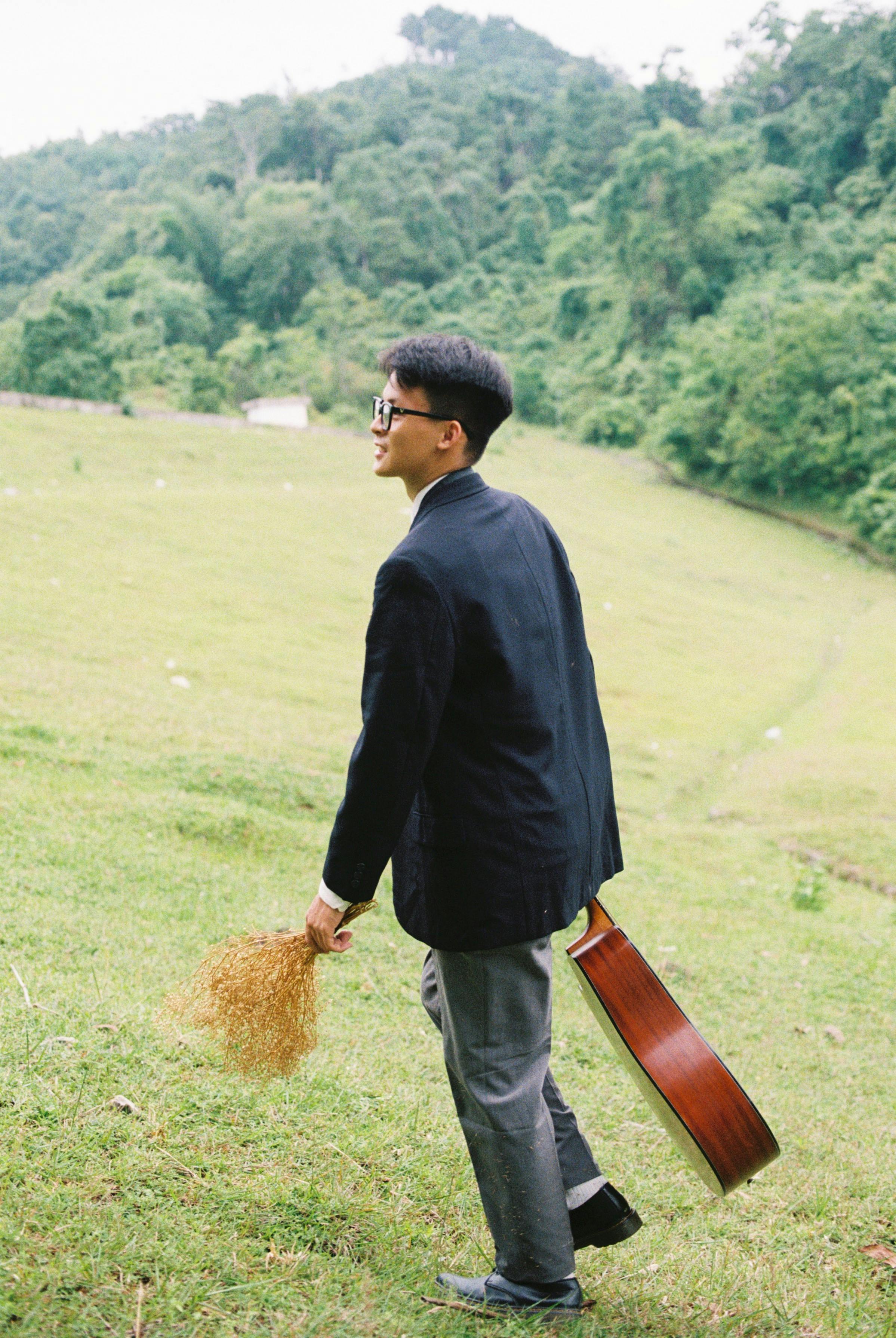Young man in countryside holding a guitar and grass, enjoying a leisurely walk in lush greenery.