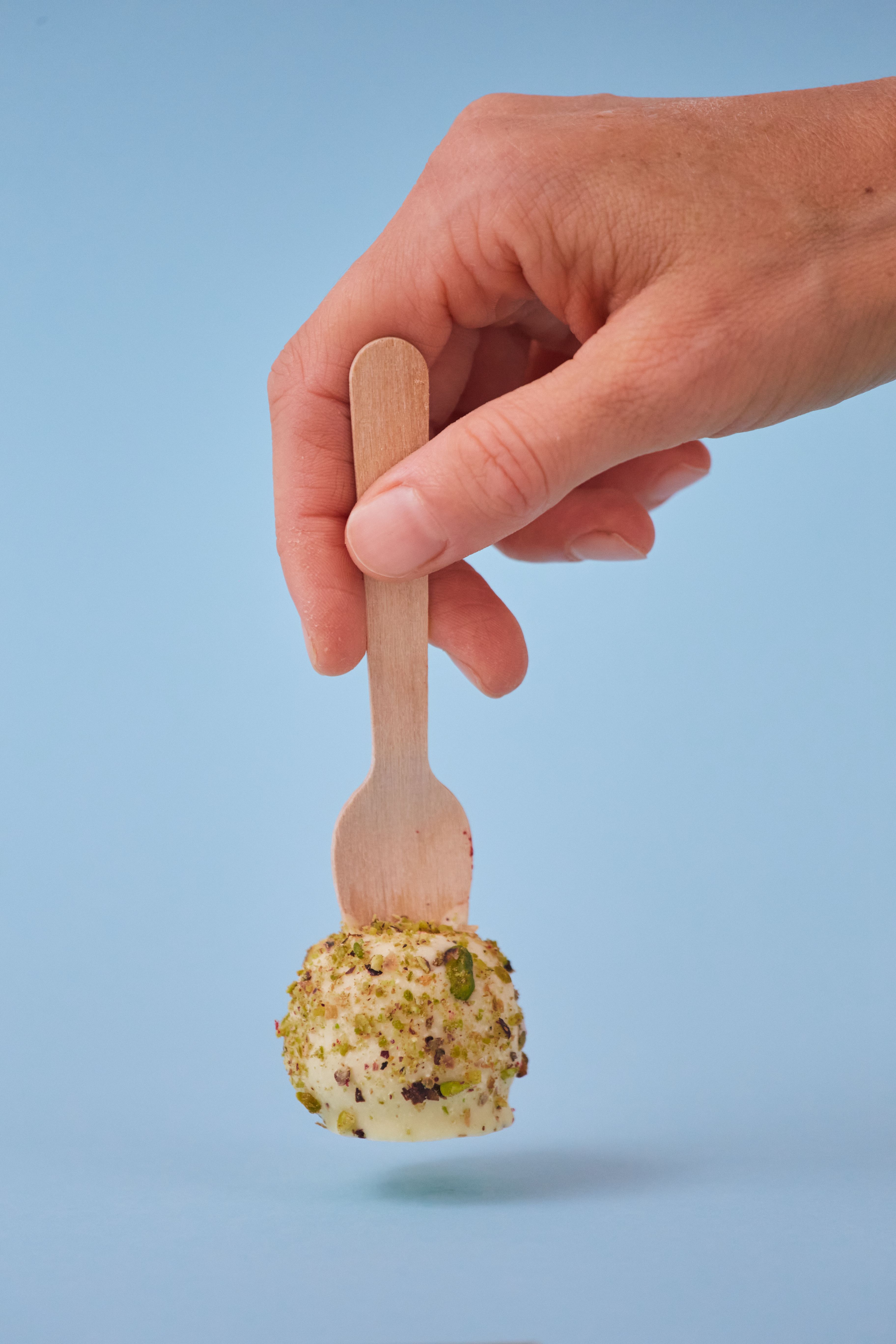 Close-up of a hand holding a pistachio-coated cake pop against a blue background.