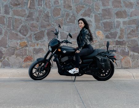 Confident woman posing on a sleek motorcycle against a stone wall in San Luis Potosí, Mexico.