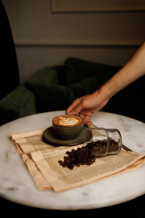 Free A hand reaches for a cappuccino on a marble table, surrounded by coffee beans and newspaper. Stock Photo