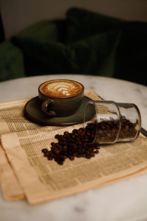 Free Elegant cappuccino with latte art alongside spilled coffee beans on a vintage newspaper table setting. Stock Photo