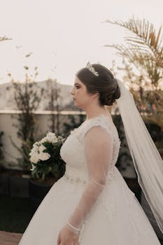 Profile of a bride holding a bouquet, wearing a lace gown and tiara outdoors.