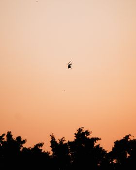 A helicopter silhouetted against an orange sky during a calming sunset, flying above treetops.