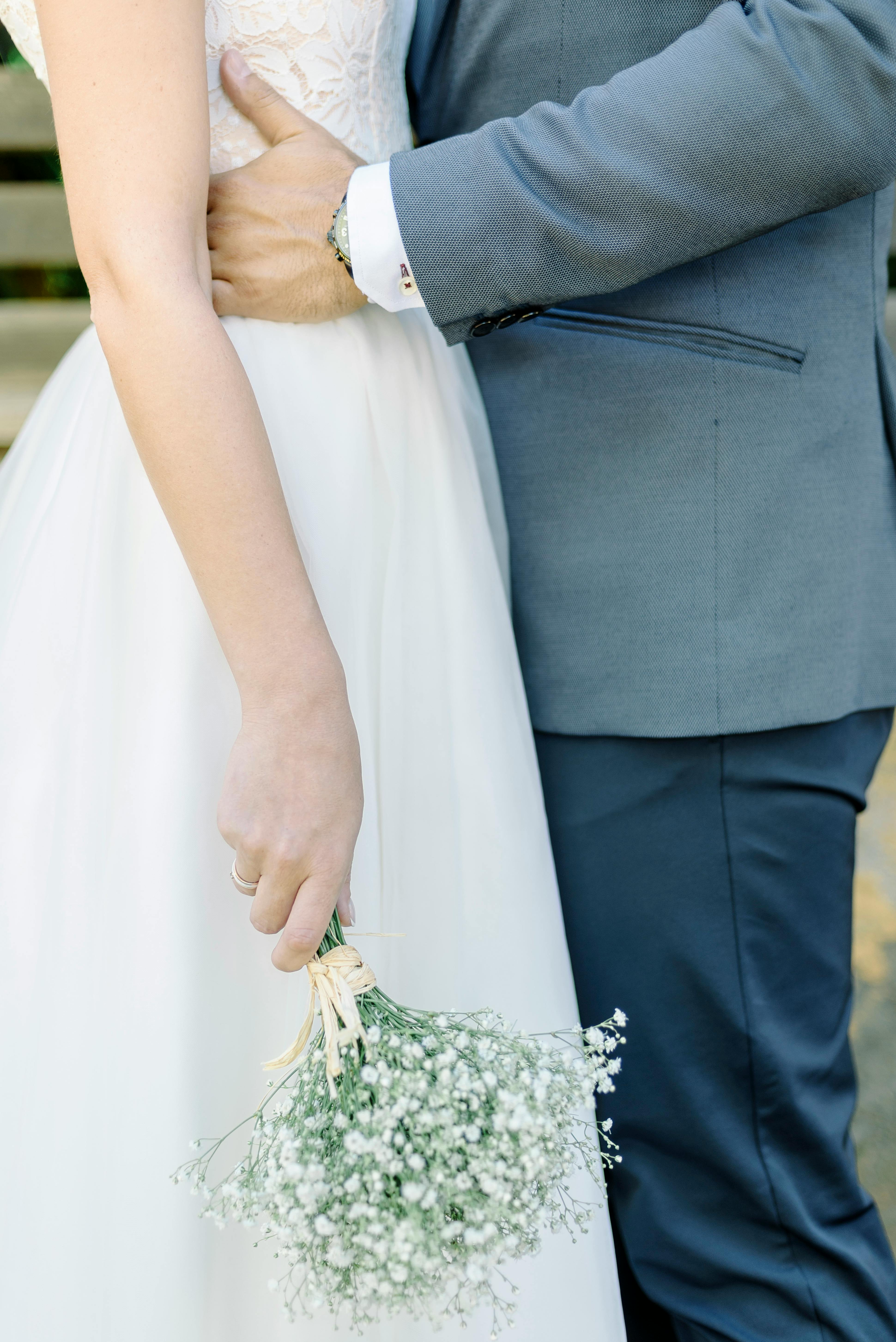 Bride and groom embrace with delicate bouquet in hand, showcasing romantic wedding attire.