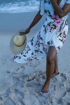 A young woman in a floral dress walks barefoot on a sandy beach in Lagos, Nigeria, carrying a straw hat.
