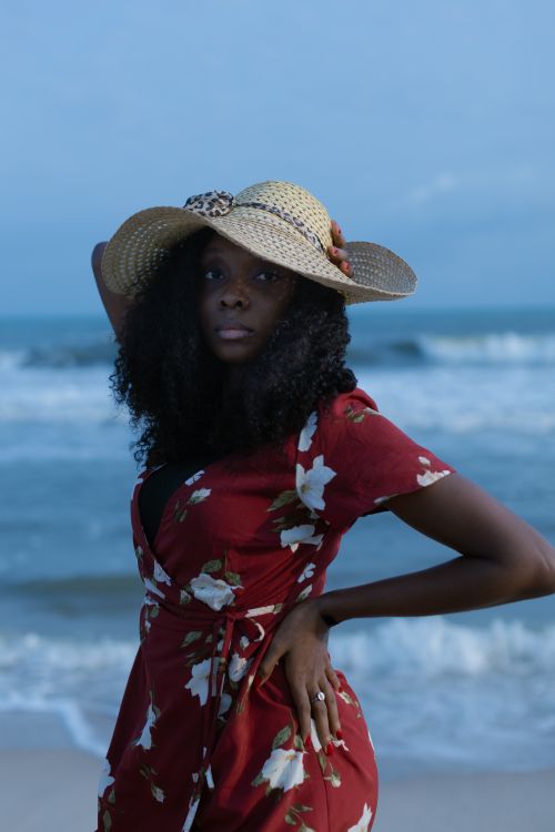 Free Stylish woman in floral dress and hat poses on Lagos beach with ocean waves in the background. Stock Photo