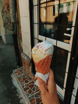 A hand holds a melting ice cream cone on a sunny day by a window in an urban setting.