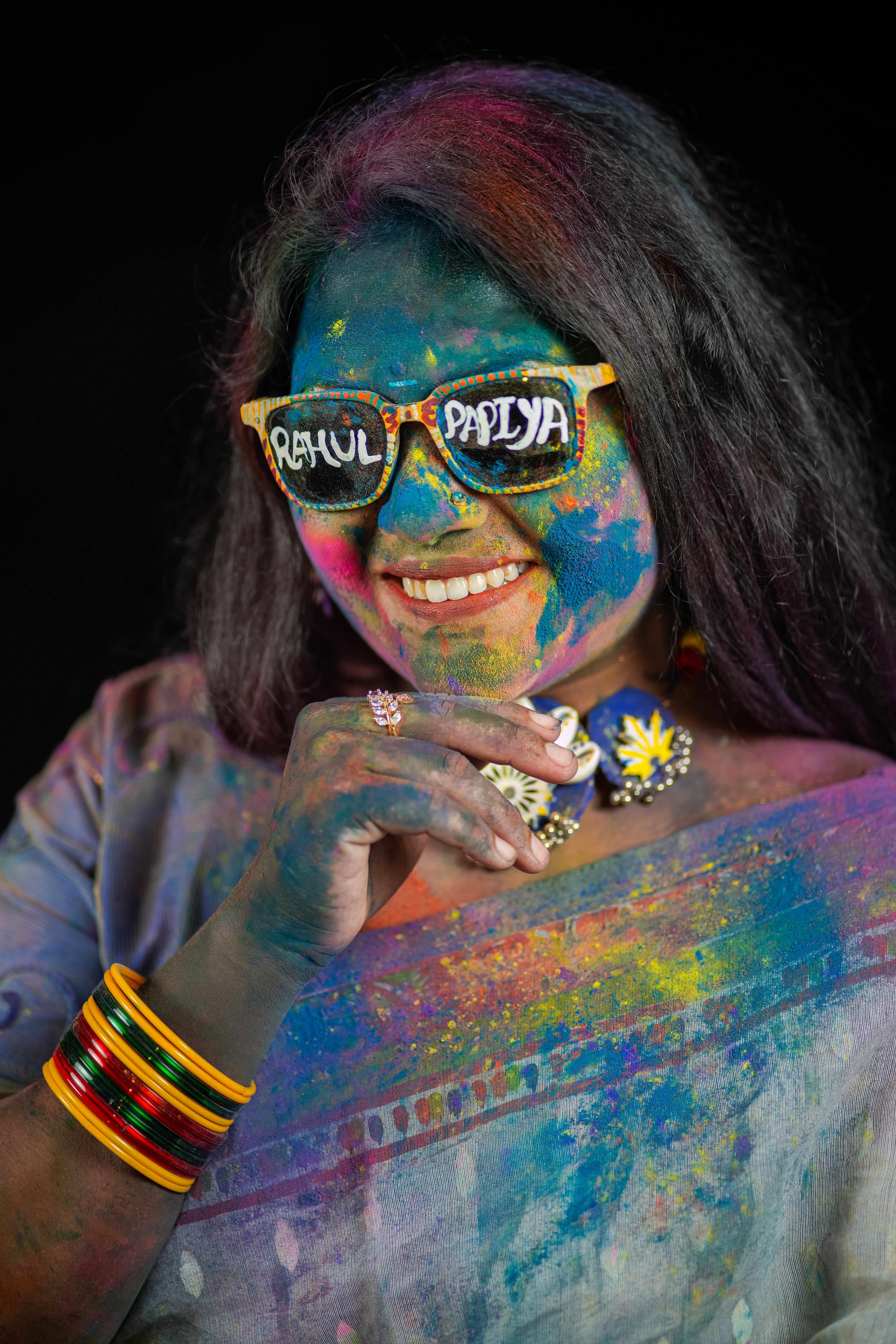 Portrait of a woman covered in vibrant Holi festival colors, wearing unique glasses and traditional jewelry.