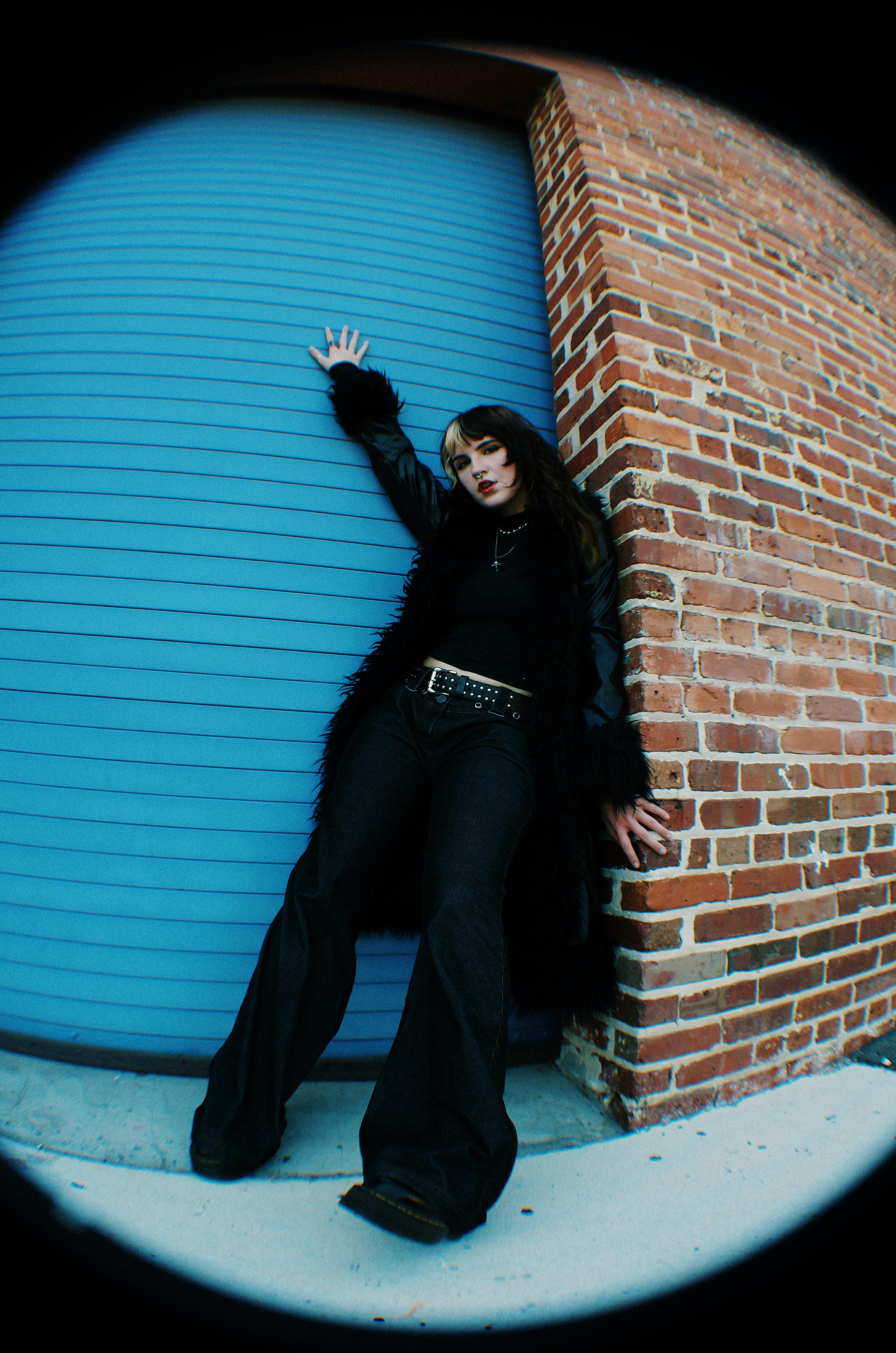 Trendy woman in black outfit leans against brick wall, exuding urban style.