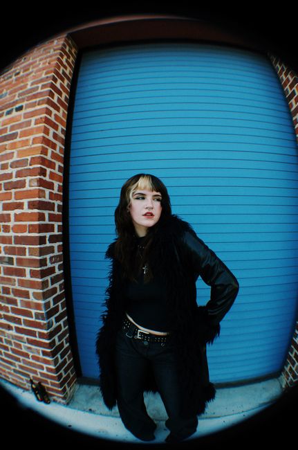 Stylish woman in black outfit poses against blue garage door with brick wall.