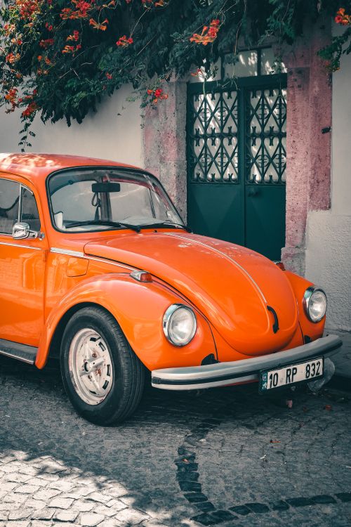 Free Classic orange Beetle car on a cobblestone street with floral backdrop. Nostalgic travel vibes. Stock Photo