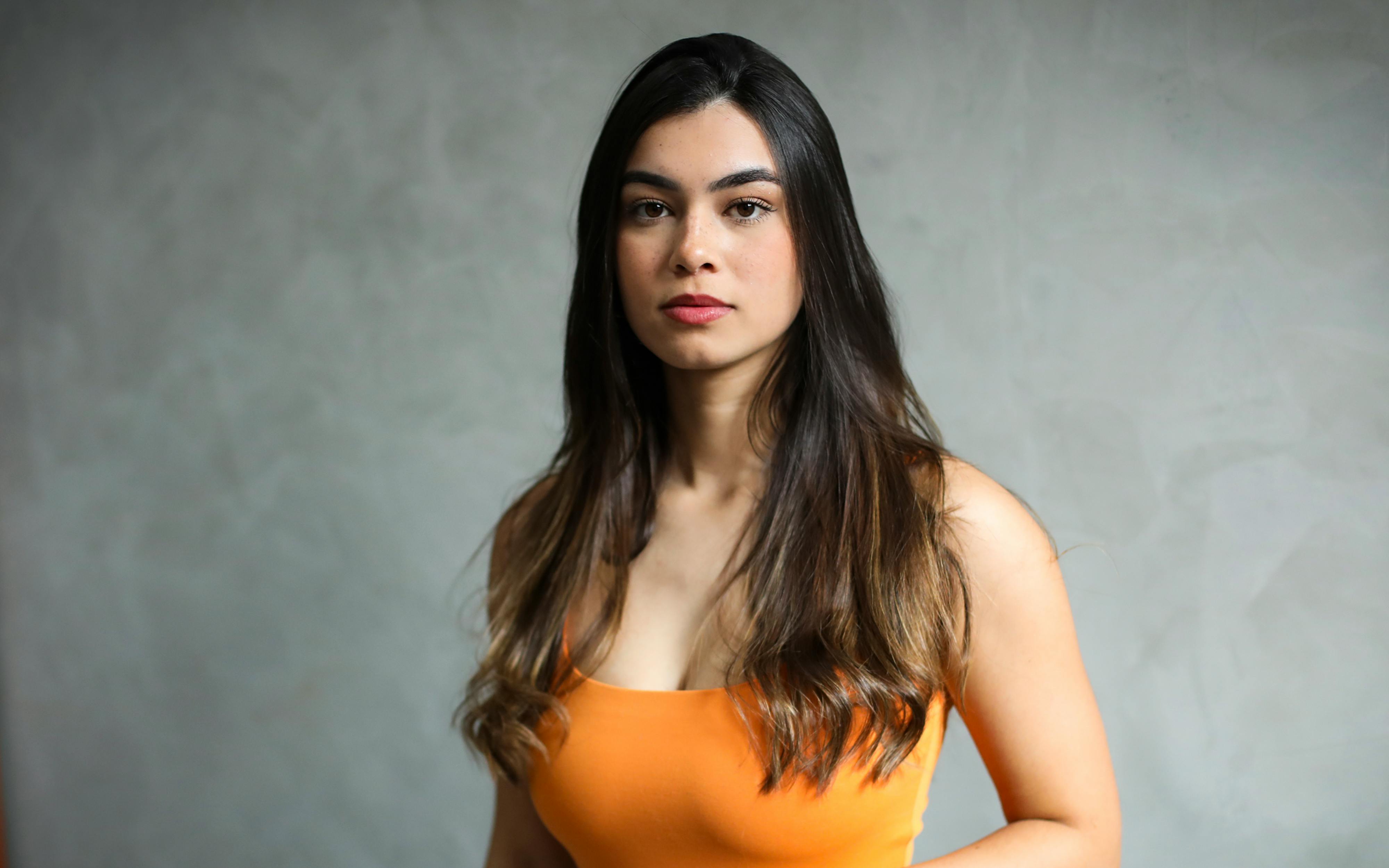 Free Elegant portrait of a young woman with long, dark hair in a vibrant orange top against a neutral background. Stock Photo