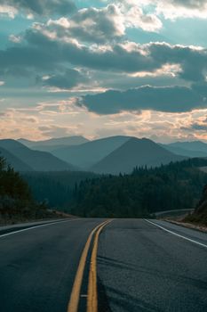 A picturesque view of a highway leading into rolling hills and mountains under a cloudy sunset sky in Kananaskis, Canada.