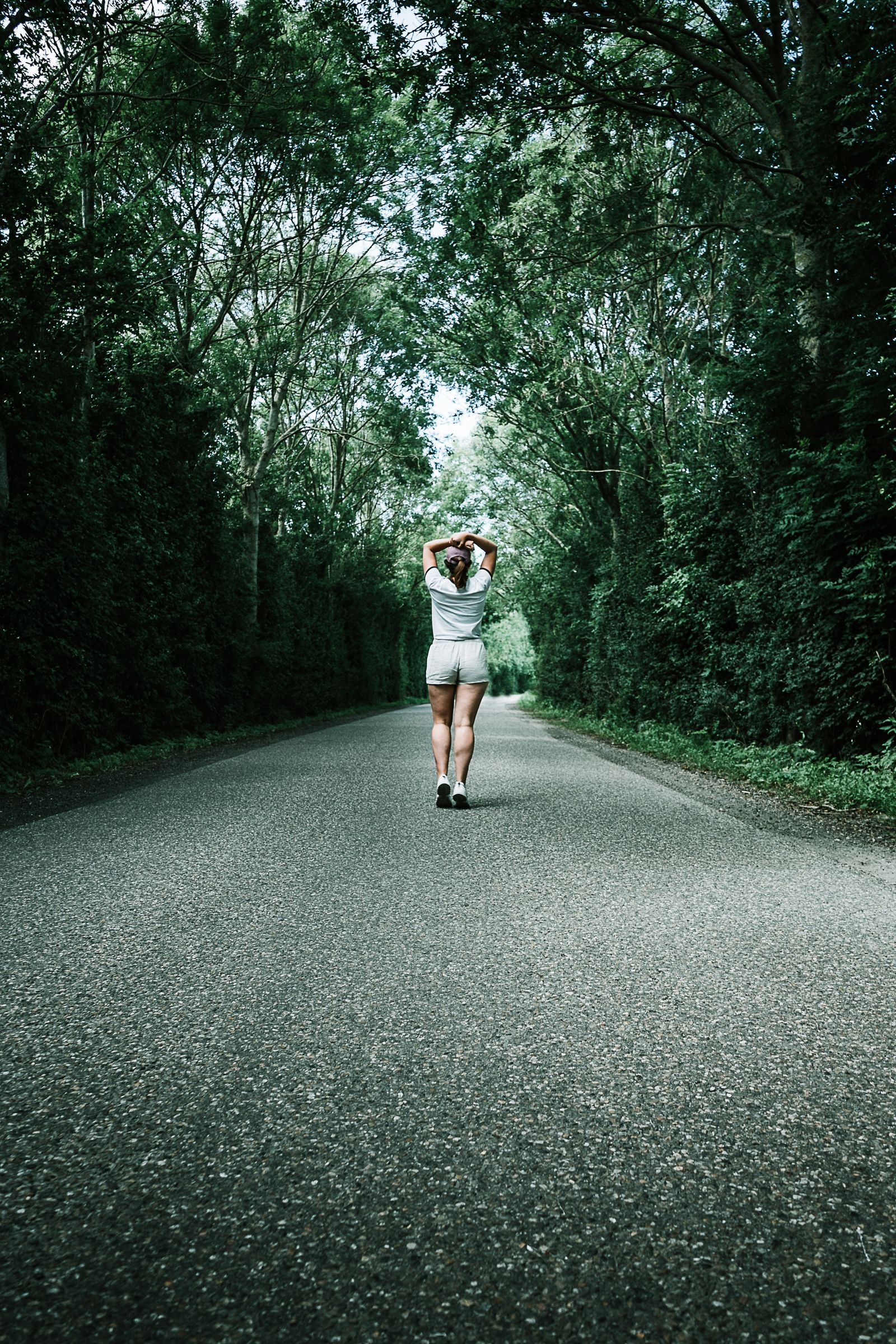 A woman in a white T-shirt walking on a quiet tree-lined road, the kind of easy after-meal walk that eases luteal-phase bloat