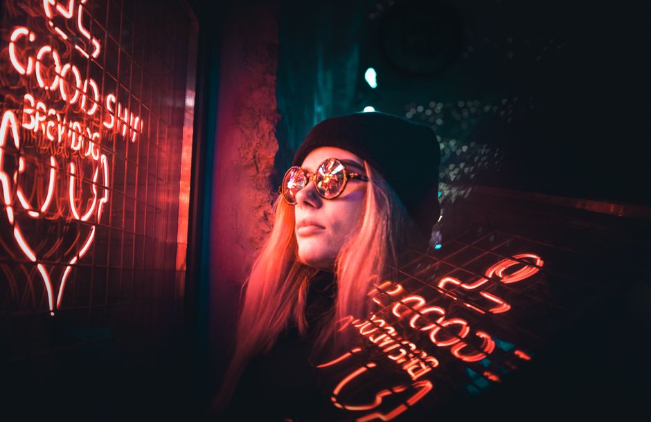 A stylish woman in sunglasses is illuminated by neon signs, creating a vibrant night scene.