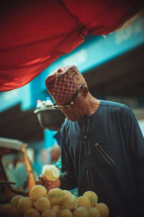 Free A man in traditional attire selects fruit at a vibrant Nigerian market under a red canopy. Stock Photo